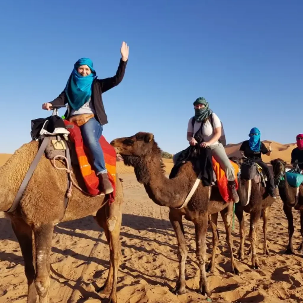 Tourists riding camels in the Moroccan desert, wearing colorful scarves and enjoying the desert scenery.